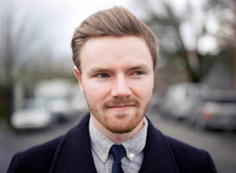 A young man with light skin, short brown hair, and a trimmed beard stands outdoors wearing a dark coat, gray shirt, and navy tie, with a slight smile. The background is blurred with trees and parked cars.