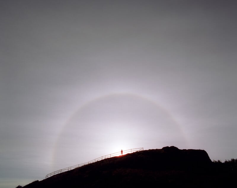A person stands on a hilltop silhouetted against the sky, with a large, faint halo surrounding the sun behind them. A fence lines the path along the ridge.