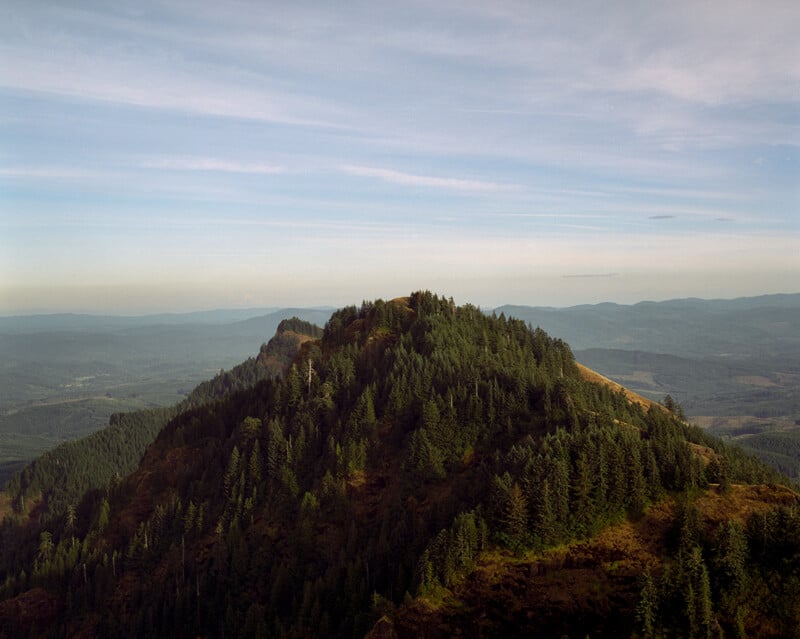 A forested mountain peak rises above rolling hills, with distant valleys and a pale blue sky stretching across the background.