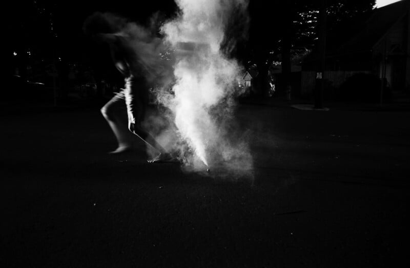 A blurry figure moves past a thick cloud of smoke on a dark, empty street at night, with houses and trees faintly visible in the background.