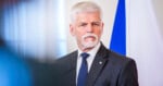 A man with white hair and a beard wearing a gray suit, white shirt, and blue striped tie stands in front of a blue and white flag, looking serious.
