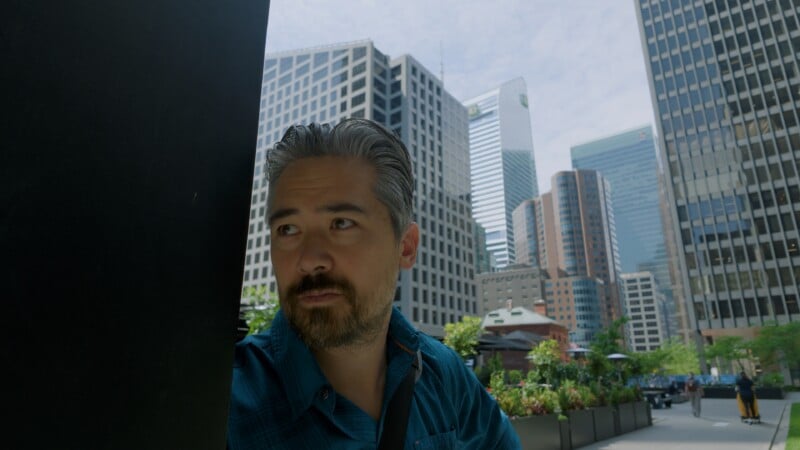 A man with dark hair and a beard, wearing a blue shirt, sits outdoors in a city plaza surrounded by tall modern skyscrapers and greenery on a sunny day.
