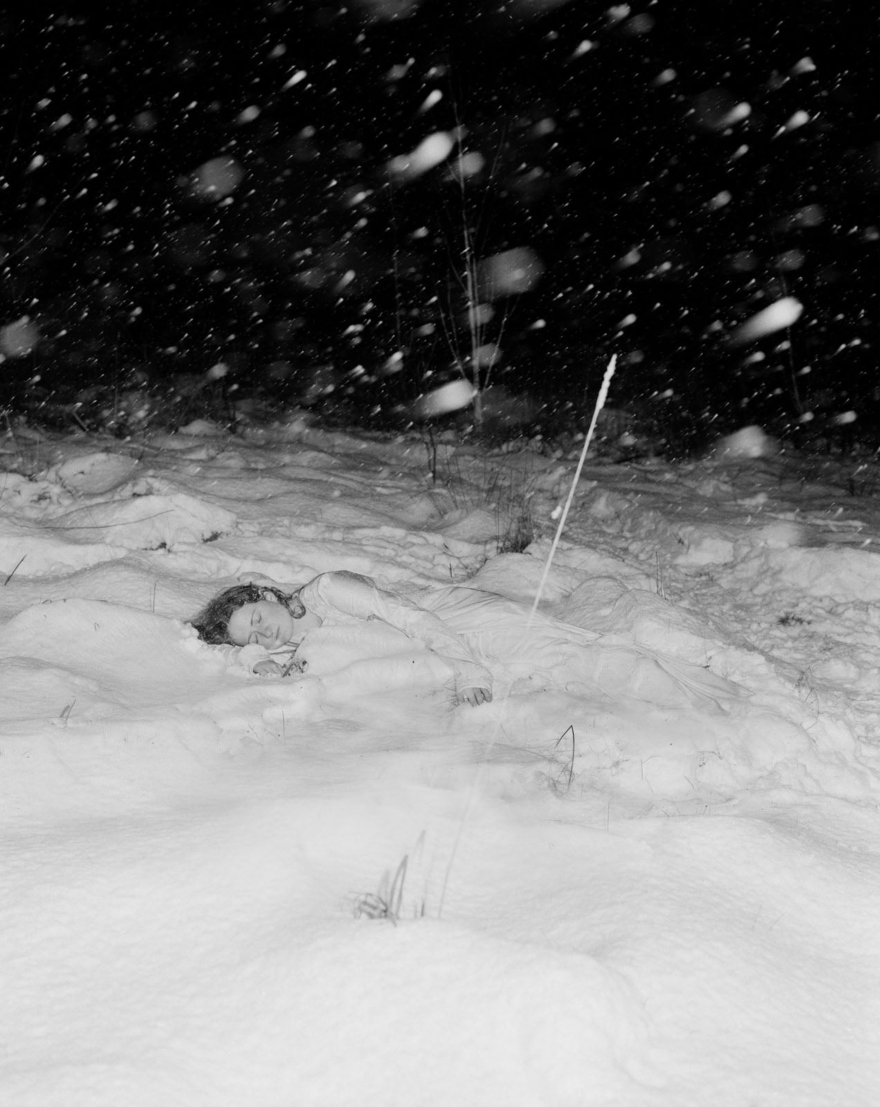 A person lies partly buried in deep snow at night, with snow falling heavily around them. Only their face and curly hair are visible above the snow in the dark, wintry scene.