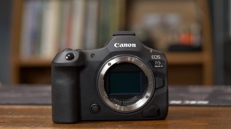 A Canon EOS R5 Mark II mirrorless camera body is placed on a wooden surface with a blurred bookshelf in the background.