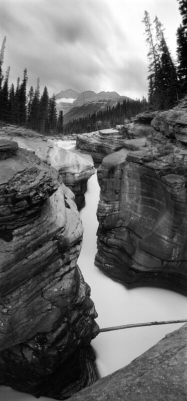 A black and white vertical shot of a narrow, winding river flowing through deep, layered rock canyons toward distant mountains.