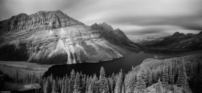 A black and white panoramic view of a dark lake nestled at the base of a large, rugged mountain range with a dense pine forest in the foreground.