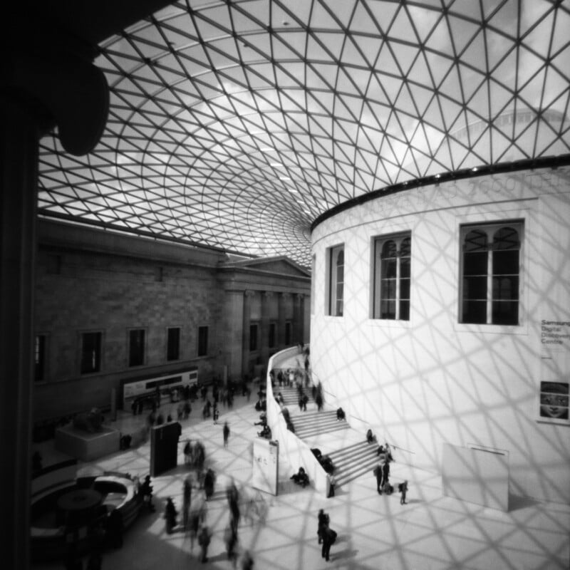 A wide view of a grand indoor atrium with a geometric glass ceiling casting shadows on the floor and walls; people walk and gather near a curved staircase and white, round building structure.