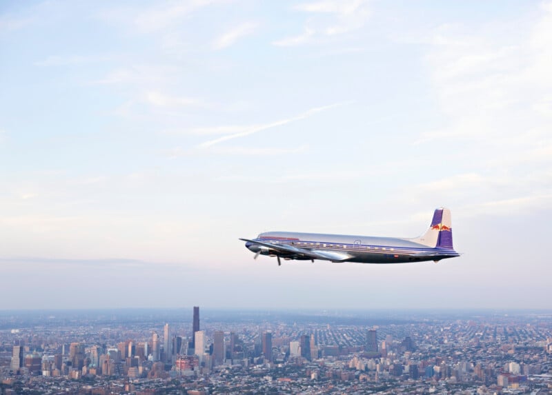 A vintage propeller airplane flies above a sprawling city skyline under a blue sky with light clouds.