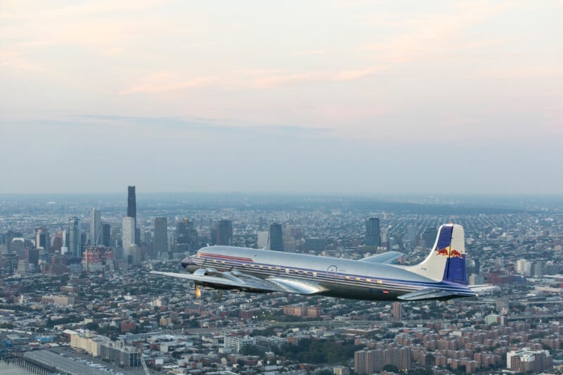 A vintage silver airplane with red and blue accents flies over a sprawling cityscape with tall buildings and a cloudy sky in the background.
