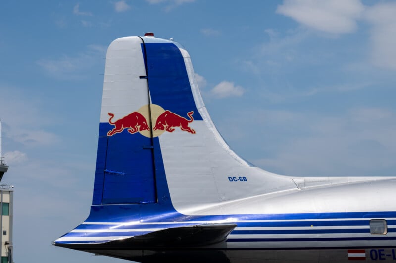 Tail section of a silver and blue aircraft with a Red Bull logo on the fin, against a partly cloudy sky. The aircraft registration "DC-6B" is visible.