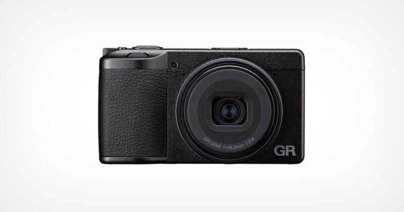 A black compact digital camera with a textured grip is shown facing forward against a plain white background. The lens is centered, and 