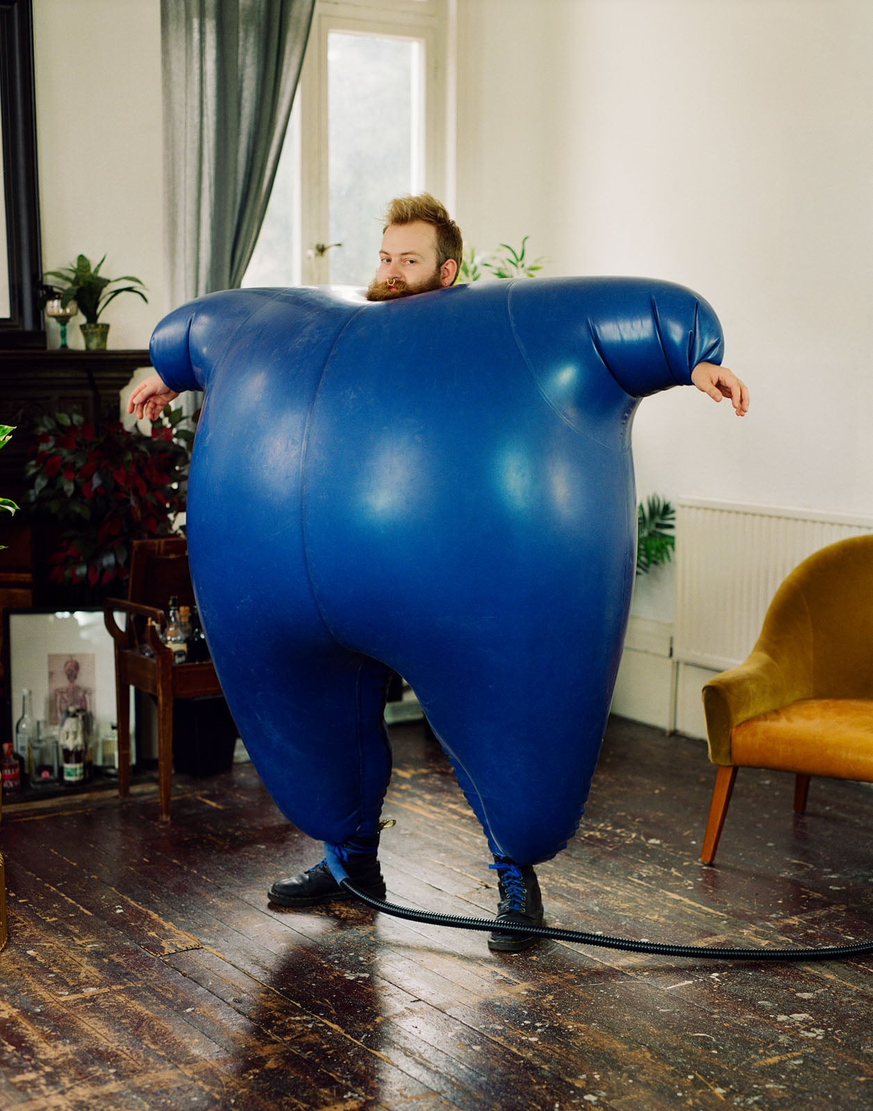 A man with a beard and a nose ring stands in a room wearing a large, inflated blue suit connected to a black hose.