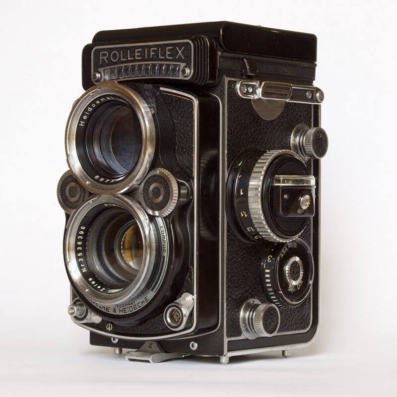 A vintage Rolleiflex twin-lens reflex camera with two large lenses, various dials, and a textured black body, displayed against a plain white background.