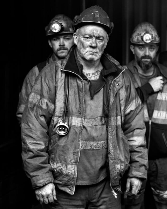 Three miners wearing helmets and work jackets, with serious expressions and dirt on their faces, stand together in a dimly lit setting. The man in front holds a flashlight and looks directly at the camera.
