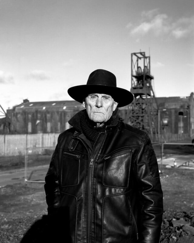 An older man in a leather jacket and wide-brimmed hat stands outdoors, looking serious. Industrial buildings and structures appear in the background under a bright sky.