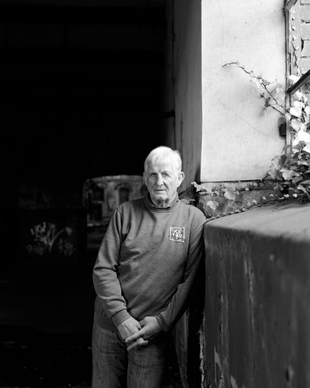 An older man with a serious expression leans against a rough, graffiti-marked wall in a dimly lit, abandoned building, with some ivy growing nearby. The image is in black and white.