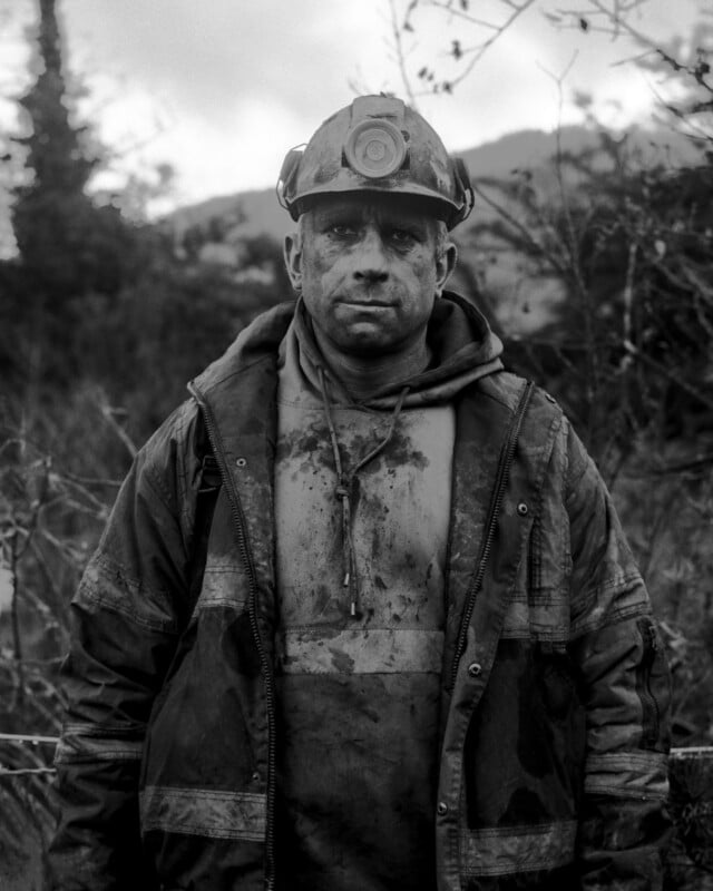 A miner wearing a headlamp and dirty work clothes stands outdoors, his face and jacket smeared with dirt. The background shows blurred vegetation and hills, suggesting a rugged, outdoor environment.