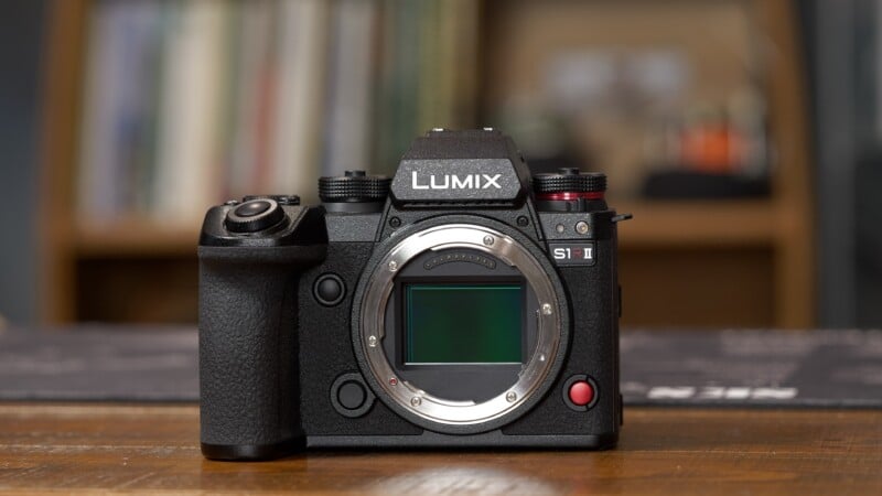 A Panasonic Lumix S1 II mirrorless camera body sits on a wooden surface with a blurred bookshelf background, showing the camera’s sensor and various controls.