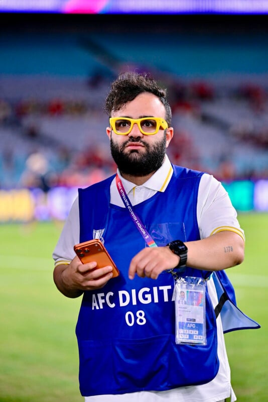 A man wearing yellow glasses, a blue "AFC Digital 08" vest, and a lanyard stands on a stadium field holding a phone, with blurred spectators in the background.