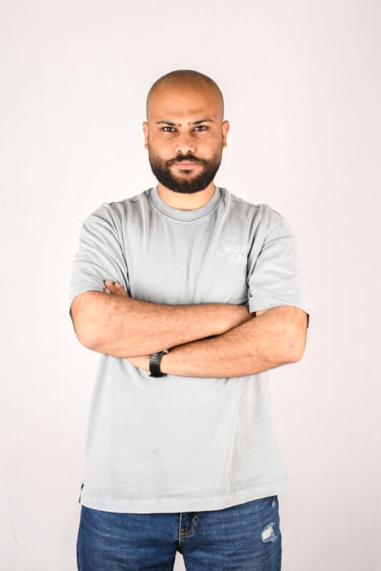 A bald man with a beard stands against a plain background, arms crossed. He wears a light gray t-shirt and blue jeans with a rip on one knee, and has a black wristwatch on his left wrist.