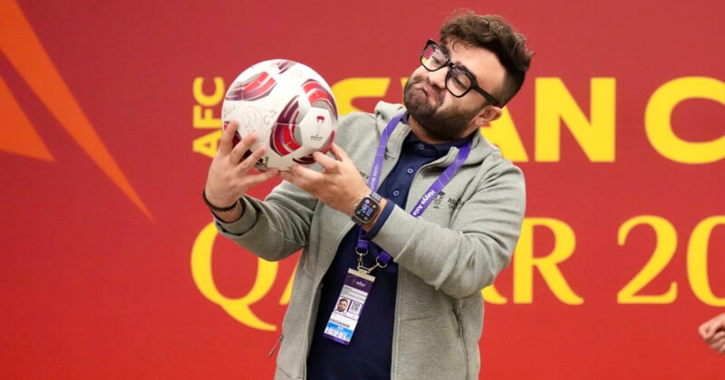 A man wearing glasses and a gray hoodie holds a soccer ball in both hands, standing in front of a red wall with large yellow text. He has an event badge and a smartwatch, and appears to be making a playful facial expression.