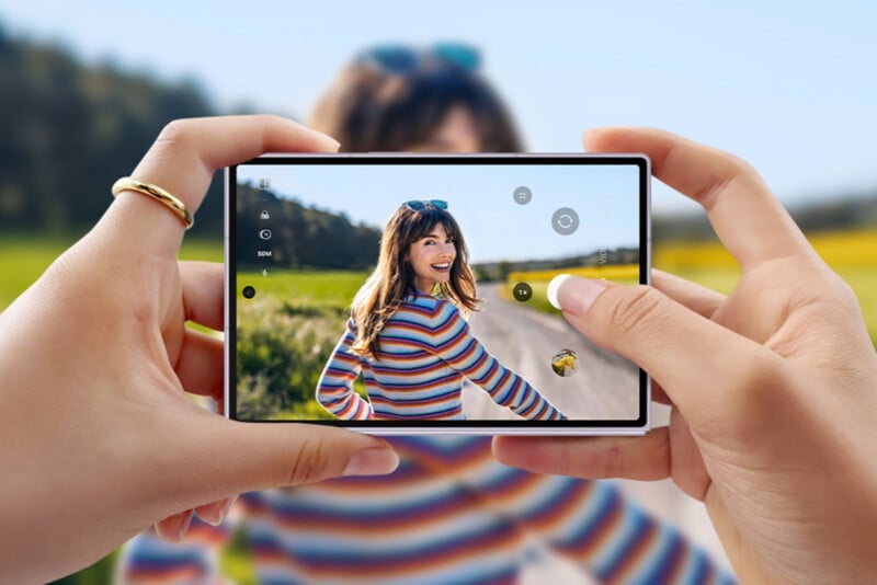 A person holds a smartphone, taking a photo of a smiling woman in a colorful striped sweater standing on a country road with greenery and blue sky in the background.