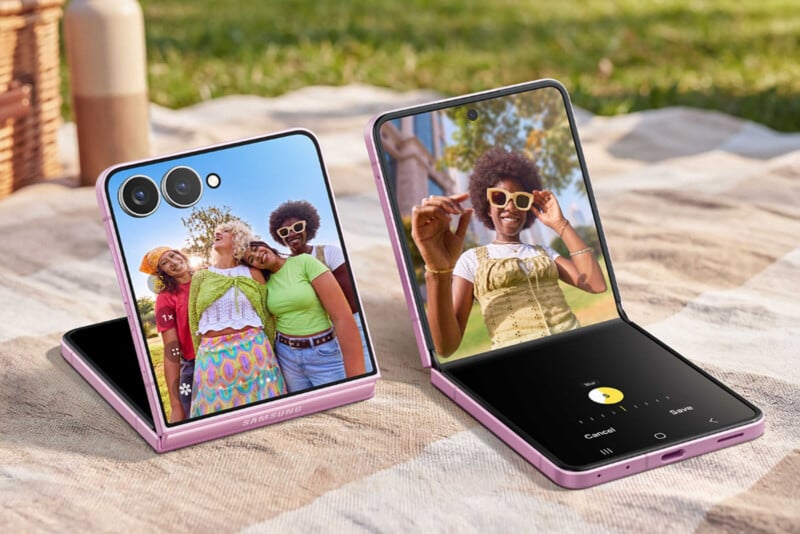 Two pink Samsung flip phones are displayed on a picnic blanket outdoors. One phone shows a selfie of three smiling friends, while the other shows a woman posing for a photo on its main screen.