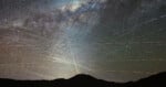 A dark silhouette of a hill is seen under a sky filled with bright star trails and the Milky Way, crisscrossed by numerous streaks of light from satellites and meteors.