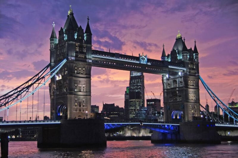 Tower Bridge in London illuminated at dusk, with a vibrant purple and pink sunset sky in the background and city buildings visible between the bridge towers.