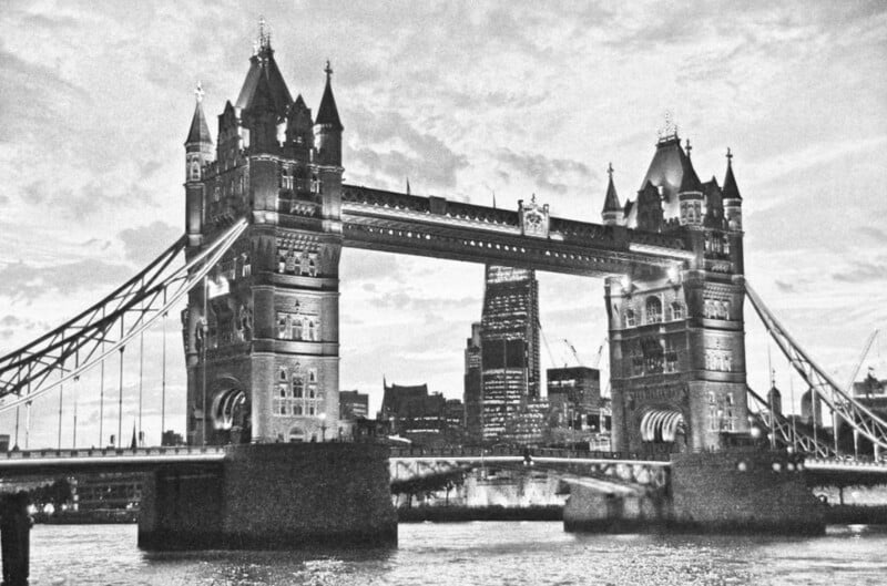 Black and white photo of London’s Tower Bridge at dusk, with its two towers lit up and the city skyline visible in the background across the River Thames.