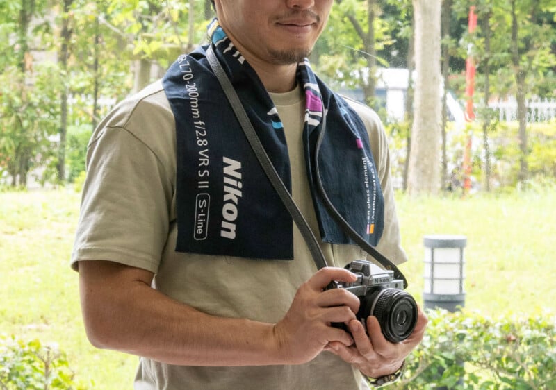 A person wearing a beige shirt and Nikon-branded towel around their neck holds a Nikon camera outdoors, with greenery and trees in the background.