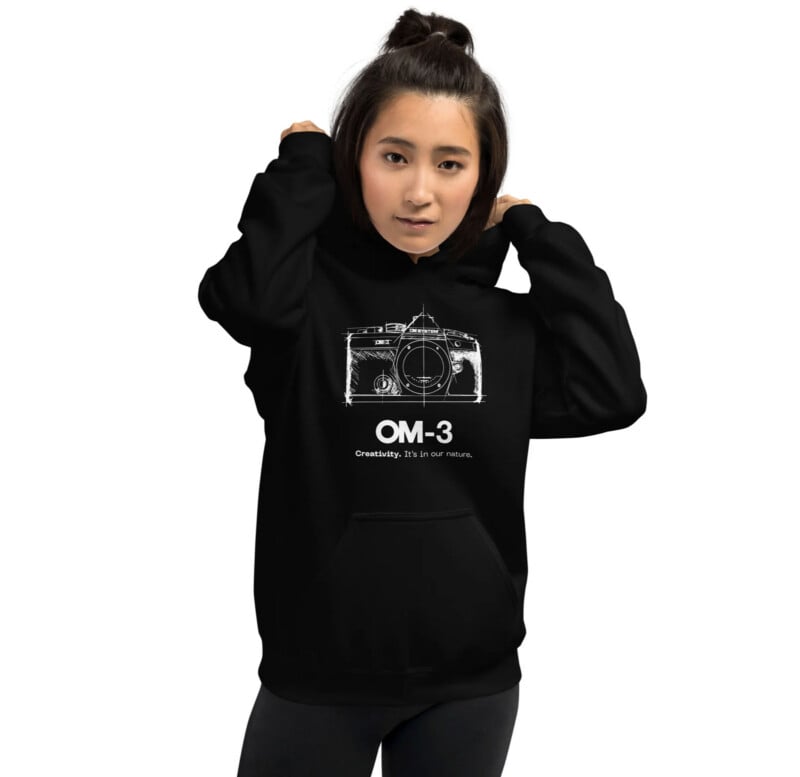 Woman wearing a black hoodie with a white camera graphic and the text “OM-3 Creativity. It’s in our nature.” printed on the front, standing against a white background.