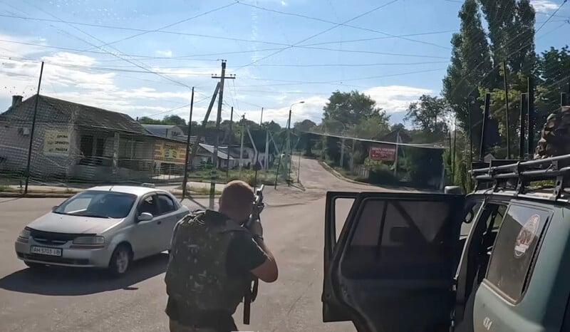 A person in military gear aims a rifle near an open vehicle door on a street. A white car is driving nearby, and buildings, trees, and power lines are visible in the background.
