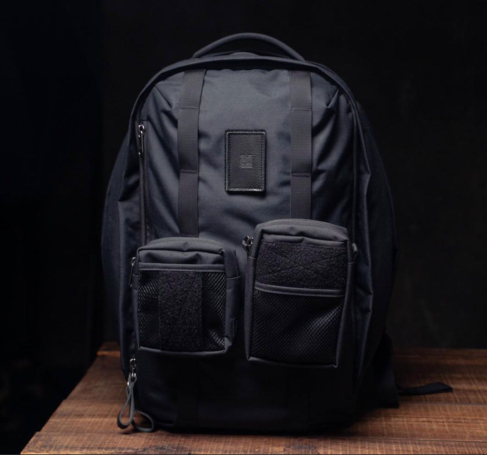 A black backpack with two front zippered pockets and mesh details sits on a wooden surface against a dark background.