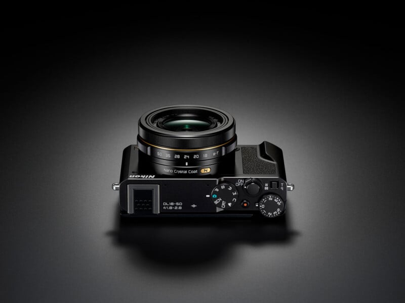 A sleek black Nikon compact camera with a lens marked “NIKKOR,” shown from above on a dark surface. The top dials, lens, and buttons are clearly visible.