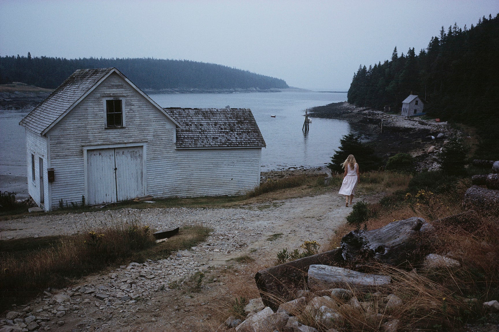 A woman in a light dress walks toward a weathered white house by a rocky shoreline, with forested land and calm water in the background under a cloudy sky.