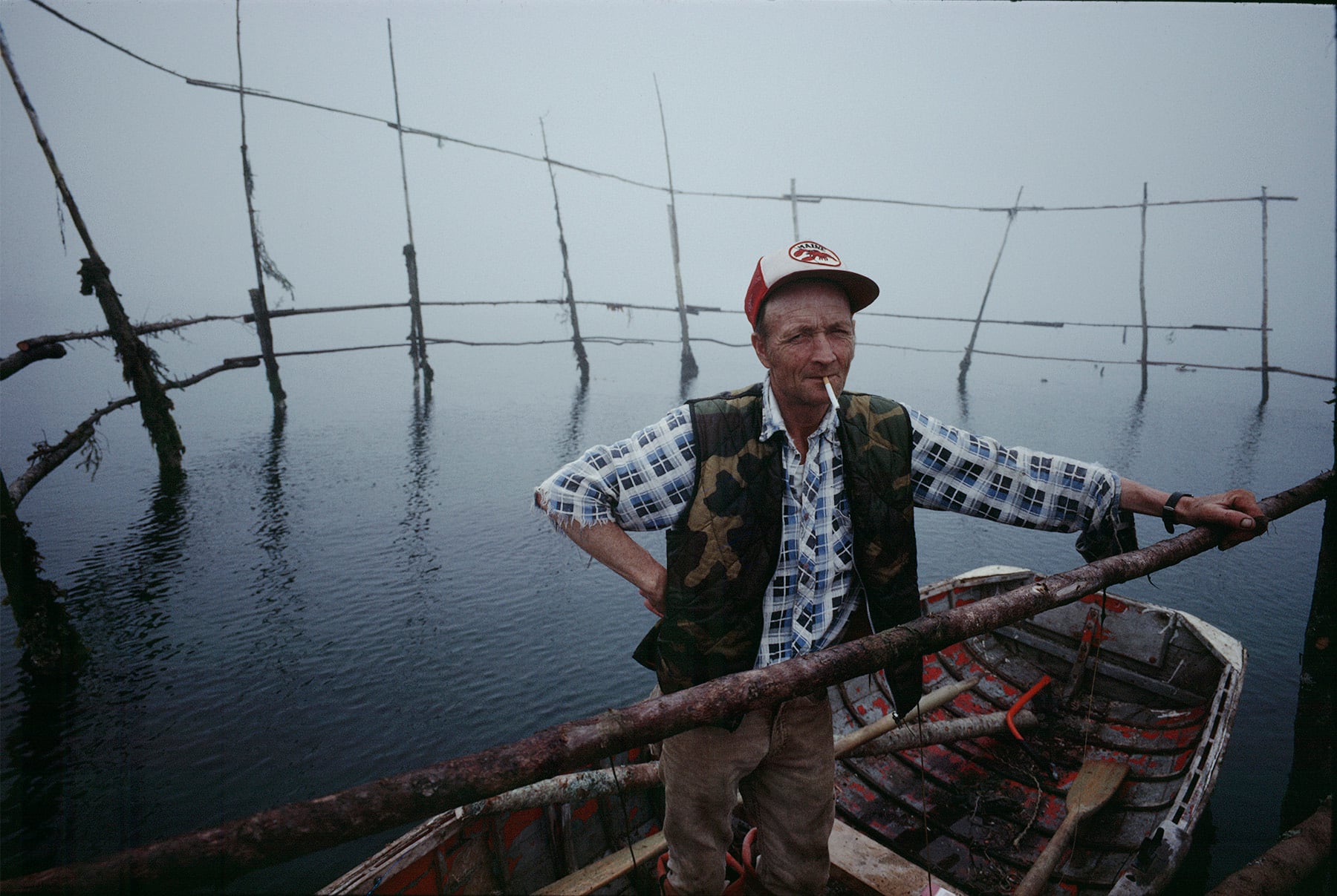 A man in a plaid shirt, camouflage vest, and cap stands in a wooden boat on a misty lake, holding onto a wooden pole, with fishing nets suspended behind him over the water.