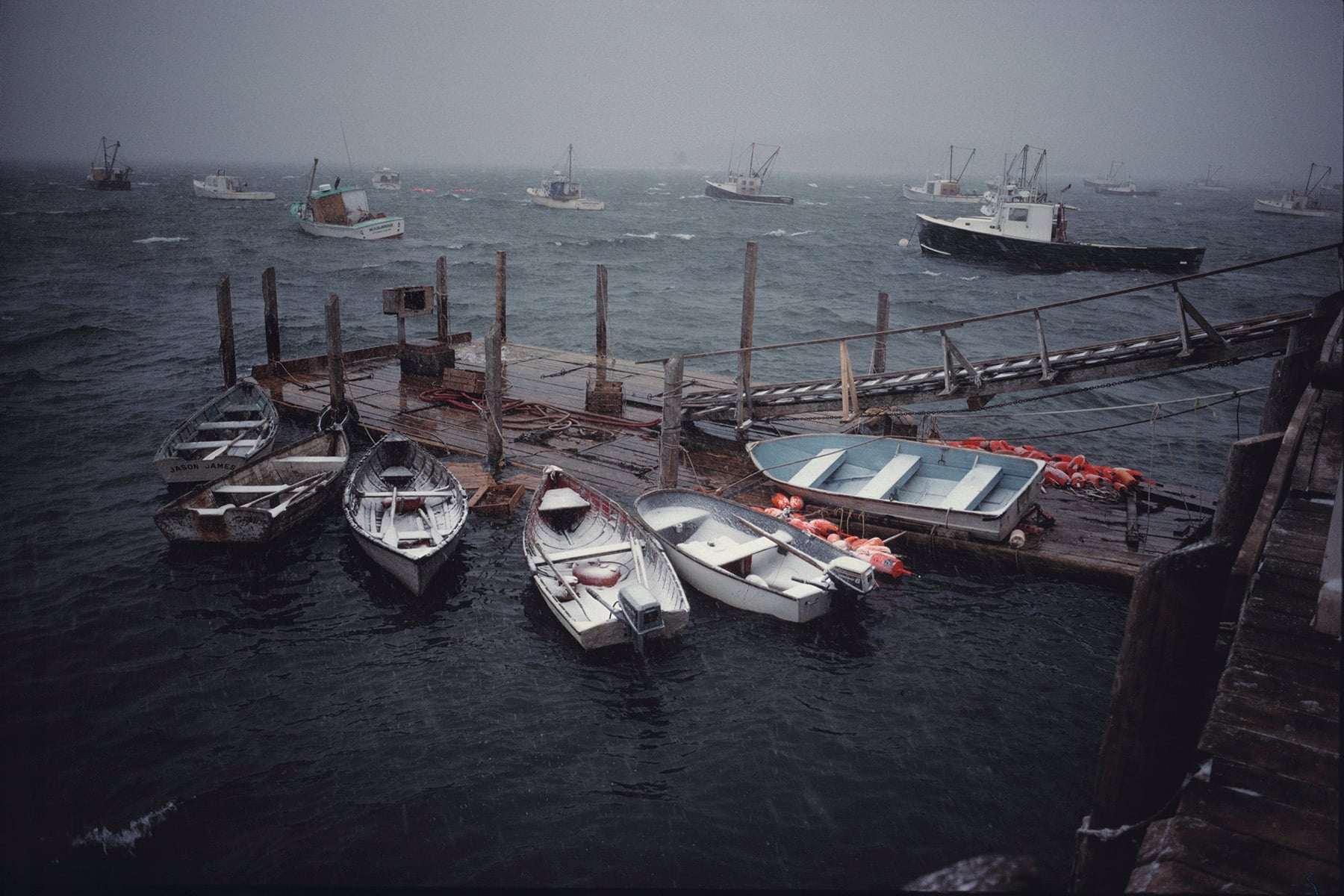 Small boats are tied to a weathered wooden dock on a choppy, overcast sea. More fishing boats are visible in the distance, shrouded in mist and rain, creating a moody, stormy atmosphere.
