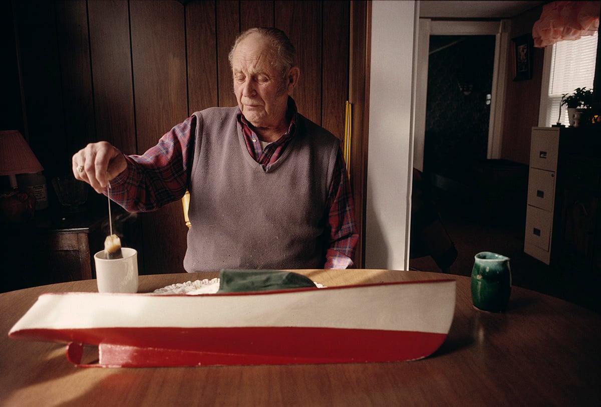 An elderly man sits at a wooden table, dipping something into a white mug. In front of him is a large model boat with red, white, and green colors. The room is warmly lit, with wooden paneling and a window in the background.