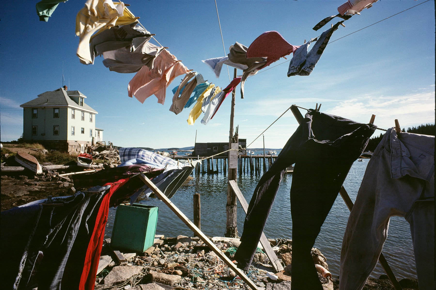 Clothes and sheets hang on lines to dry in a lakeside yard, with a pier and dock in the background. A white house stands near the water under a clear blue sky.