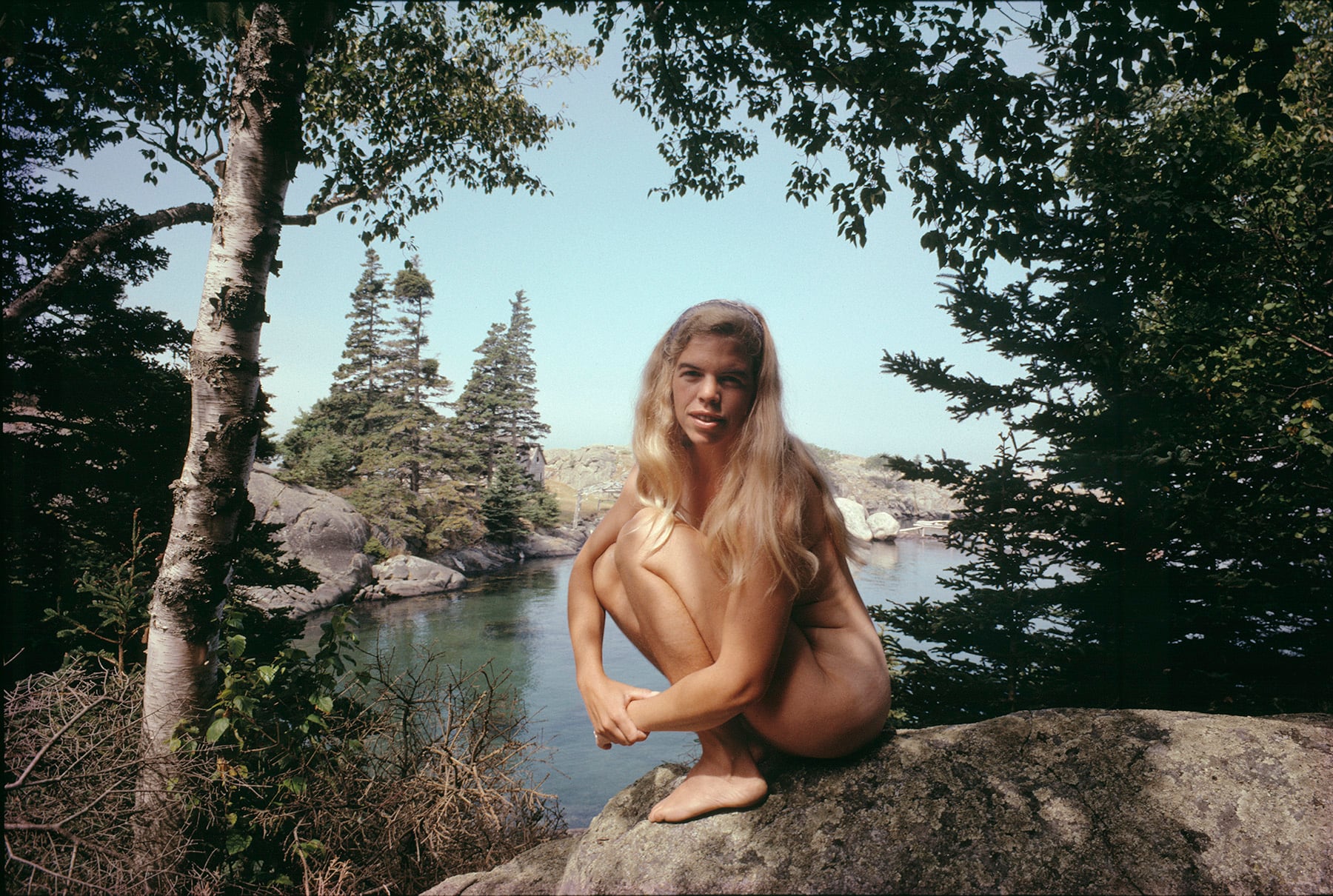A woman with long blonde hair sits nude on a rock by a lake, surrounded by trees and natural scenery on a sunny day.