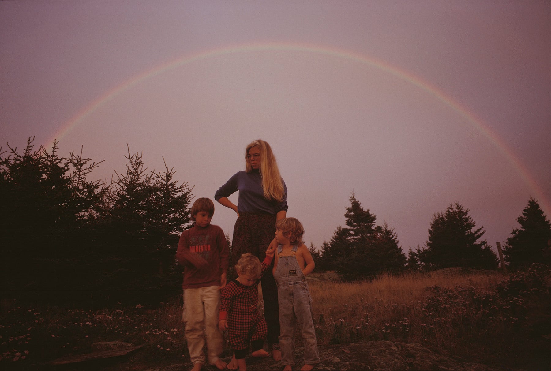 A woman with long blonde hair stands outdoors with three young children in front of her, under a rainbow arcing across a cloudy sky and surrounded by trees and grassy landscape at dusk.