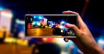 A hand holds a smartphone, capturing a photo of emergency vehicles with flashing lights at night, possibly at an accident scene. The phone screen shows a clearer image than the blurred background.