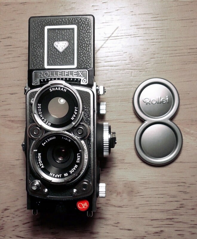 A vintage Rolleiflex twin-lens reflex camera is placed on a wooden surface next to a metallic Rollei lens cap. The camera displays intricate dials and lenses, with a textured pattern on its upper section.
