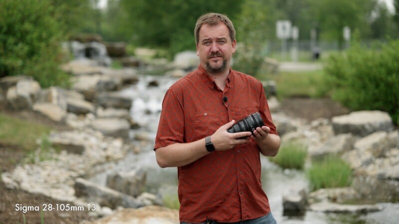 A man in a red shirt stands outdoors by a rocky creek, holding a camera lens. Trees and greenery are in the background. Text reads "Sigma 28-105mm T3" in the lower left corner.