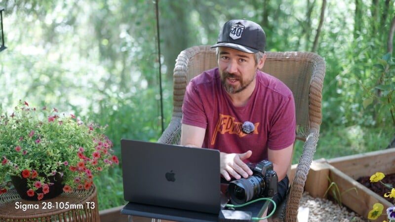 A man in a red shirt and cap sits outdoors on a wicker chair, using a laptop. He holds a camera and is surrounded by greenery and flowers. Text in the corner reads "Sigma 28-105mm T3".