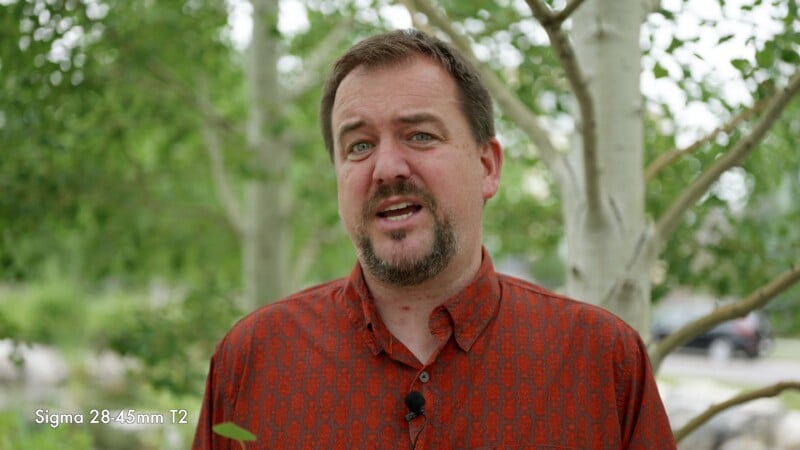 A man with a short beard and mustache, wearing a red patterned shirt, stands outdoors in front of green trees. A microphone is clipped to his shirt. Text reads, "Sigma 28-45mm T2" in the lower left corner.