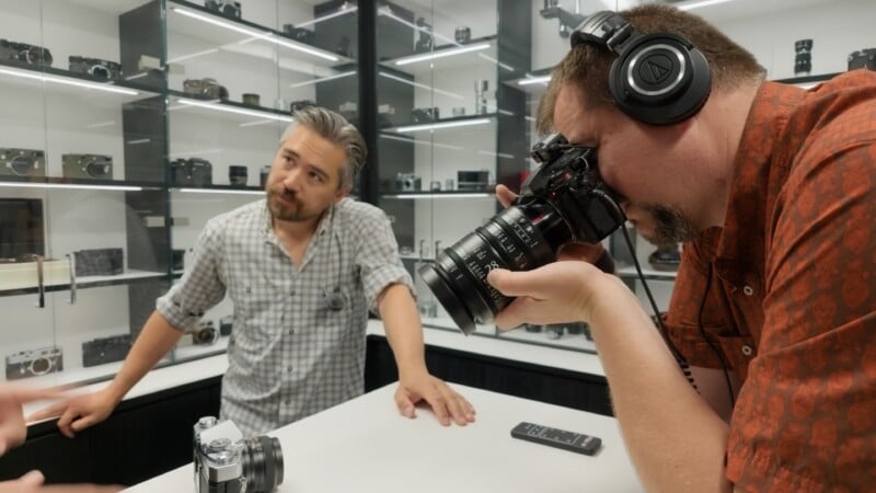 Two men in a camera store; one in headphones takes a photo with a DSLR, while the other leans on a counter. Various cameras and lenses are displayed on shelves behind them.