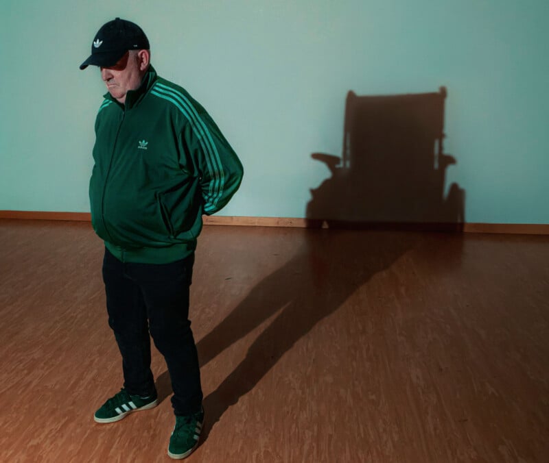 A man in a green tracksuit stands on a wooden floor, casting a shadow that resembles a wheelchair against a blank wall.