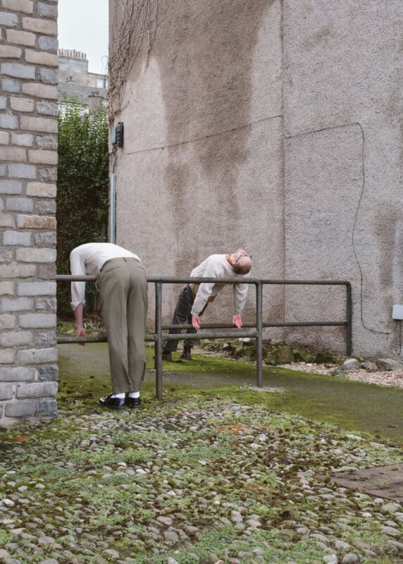 Two people bend forward over a metal railing in an outdoor alleyway between buildings, with their arms hanging down and heads lowered. Moss and stones cover the ground around them.